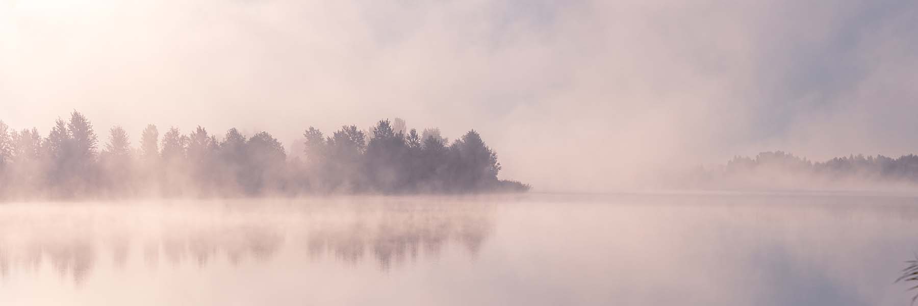 Fog rising up off of the lake casts a pinkish brown hue. A wooded area extends beyond the far side of the lake.